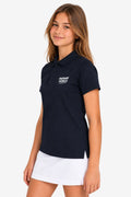 Polo technique fille bleu marine, col polo, logo poitrine, porté avec jupe blanche.