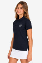 Polo technique fille bleu marine, col polo, logo poitrine, porté avec jupe blanche.
